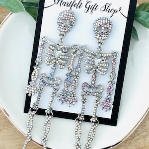 May include: A pair of silver-toned skeleton earrings featuring iridescent rhinestones and a clear teardrop-shaped center stone. The earrings are displayed on a white card with a black border that reads Hartfelt Gift Shop in black script.