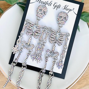 May include: A pair of dangling skeleton earrings featuring iridescent rhinestones set in silver-tone metal. Each earring depicts a full articulated skeleton figure with a clear teardrop-shaped stone at the chest, displayed on a Hartfelt Gift Shop card.
