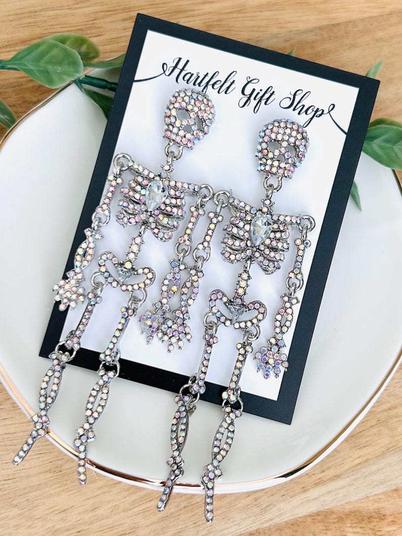 May include: A pair of dangling skeleton earrings featuring iridescent rhinestones set in silver-tone metal. Each earring depicts a full articulated skeleton figure with a clear teardrop-shaped stone at the chest, displayed on a Hartfelt Gift Shop card.