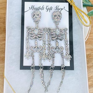 May include: A pair of silver-toned skeleton earrings featuring iridescent rhinestones and a central clear teardrop-shaped crystal on the chest. The earrings are displayed on a Hartfelt Gift Shop card inside a white gift box with gold ribbon.