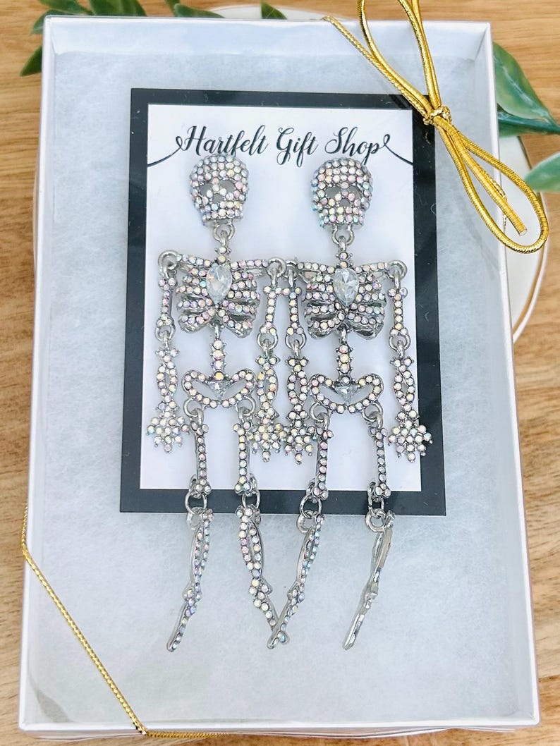 May include: A pair of silver-toned skeleton earrings featuring iridescent rhinestones and a central clear teardrop-shaped crystal on the chest. The earrings are displayed on a Hartfelt Gift Shop card inside a white gift box with gold ribbon.
