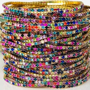 Könnte beinhalten: Ein Stapel goldfarbener Armbänder, jedes mit einer Reihe farbenfroher, funkelnder Strasssteine verziert. Die Armbänder zeigen eine lebendige Farbpalette, darunter Rosa, Blau, Grün und Gelb, die einen schillernden Effekt erzeugt. Diese Accessoires sind perfekt.