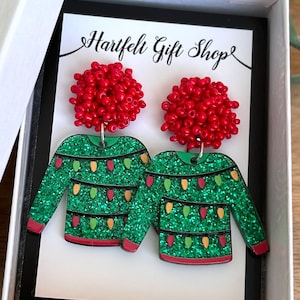 May include: A pair of festive earrings in a gift box. The earrings feature a green glitter sweater design with colorful lights and red beaded tops. The gift box has the text "Hartfelt Gift Shop" on the card.