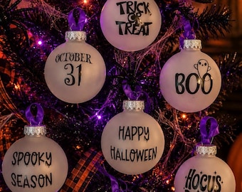 May include: A collection of round, frosted glass ornaments featuring black text and graphics. The ornaments display phrases including Trick or Treat, October 31, Boo with a ghost, Happy Halloween, Spooky Season, and Hocus Pocus, each with a purple ribbon.