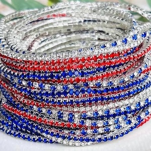 Pode incluir: Uma pilha de pulseiras prateadas adornadas com strass vermelhos, brancos e azuis. As pulseiras estão dispostas em camadas, com faixas alternadas de pedras vermelhas e azuis. Estas pulseiras são provavelmente acessórios de moda ou joalharia.