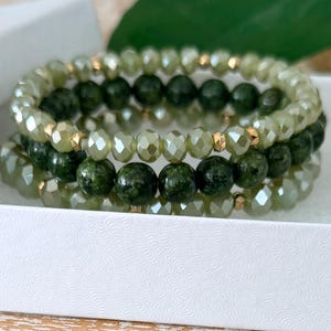 May include: Three beaded bracelets in a white box. The bracelets feature a mix of green and gold beads. The top and bottom bracelets have faceted, light green beads, while the middle bracelet has dark green, round beads. Gold-colored spacers are between the beads.