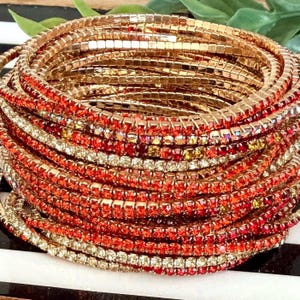 May include: A stack of multiple flexible bracelets featuring rows of small, square-cut orange and red rhinestones set in a gold-toned metal frame. The bracelets are arranged in a circular formation, showcasing a vibrant, reflective, and metallic finish.