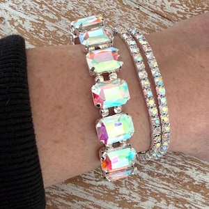 May include: Two bracelets on a wrist. One bracelet features large, rectangular, iridescent crystals set in silver-toned metal. The other is a delicate tennis bracelet with small, clear crystals. The bracelets are displayed on a light-toned wooden surface.