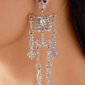 May include: A dangling earring shaped like a full human skeleton, crafted from silver-toned metal and covered in iridescent, multi-colored rhinestones. The skeleton figure features a skull stud, a ribcage, and articulated arms and legs.