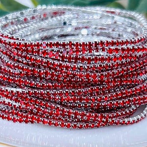 May include: A stack of red and silver rhinestone bracelets. The bracelets are made of small, sparkling red rhinestones set in a silver metal band. The bracelets are stacked on a white plate, creating a layered effect. The overall theme is jewelry.