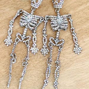 May include: A pair of dangling earrings shaped like skeletons, covered in small iridescent rhinestones. Each skeleton features a larger, clear, teardrop-shaped rhinestone at the center of the chest and a smaller one at the pelvic area.