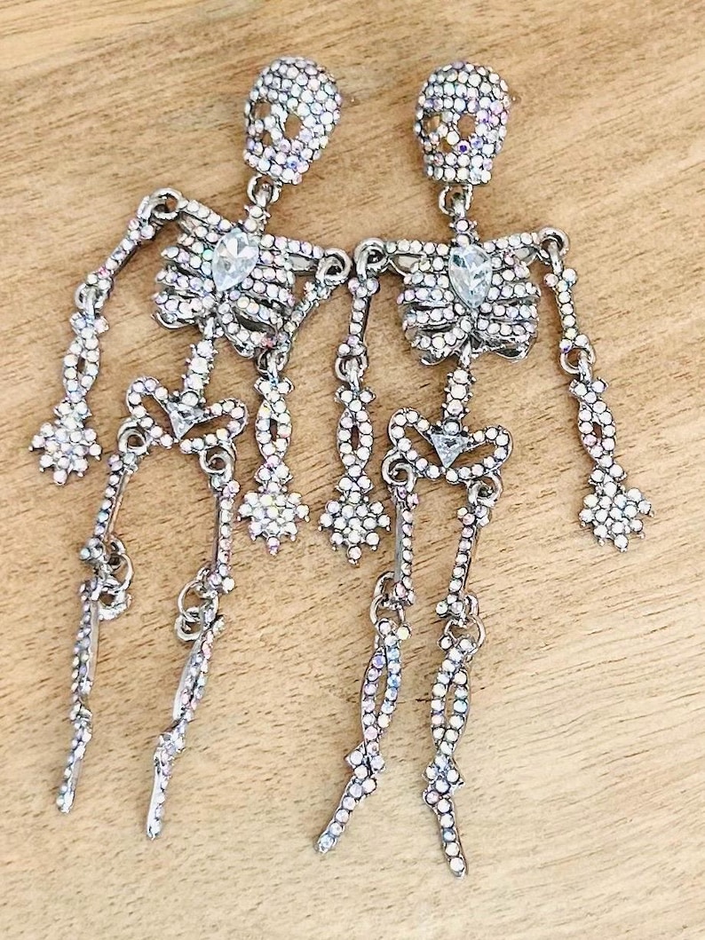 May include: A pair of dangling earrings shaped like skeletons, covered in small iridescent rhinestones. Each skeleton features a larger, clear, teardrop-shaped rhinestone at the center of the chest and a smaller one at the pelvic area.