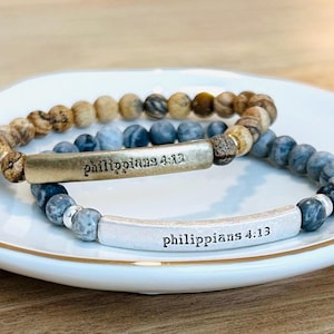 May include: Two beaded bracelets with the text "Philippians 4:13" on a white plate. One bracelet features brown and tan beads, while the other has blue and gray beads. The bracelets are made of natural stone.