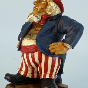 May include: A figurine of Uncle Sam, a patriotic figure, stands on a brown base. He wears a blue coat, red and white striped pants, and a matching top hat. His face has a stern expression, with a white beard and a red bow tie.