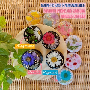 May include: A collection of round phone grips with dried flowers and gold leaf encased in clear resin. Some grips are labeled "Magnetic" and "Regular Pop-out". The text in the image reads "MAGNETIC BASE IS NOW AVAILABLE FOR BOTH IPHONE AND SUMSUNG WIRELESS CHARGER."