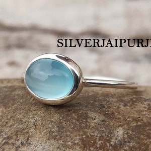 Op de afbeelding: Een zilveren ring met een lichtblauwe ovale edelsteen. De ring ligt op een bruin, getextureerd oppervlak.