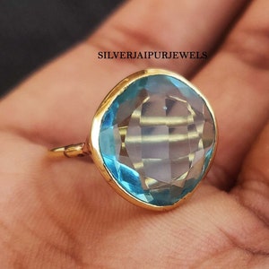 May include: A gold ring featuring a large, faceted, light blue gemstone. The gemstone is cushion-cut and set in a gold bezel. The ring is displayed on a hand, showcasing its color and design.