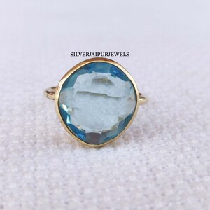 May include: A gold ring with a large, light blue gemstone. The gemstone is cushion-cut and set in a gold bezel. The ring is on a white, textured surface. The text "SILVERJAIPURJEWELS" is visible.