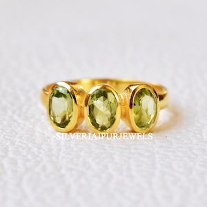 Peut inclure: Une bague en or avec trois pierres précieuses vertes de forme ovale. La bague est sur un fond blanc.