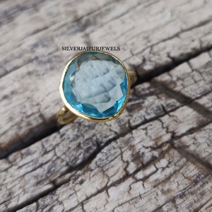 May include: A gold ring with a large, light blue gemstone. The gemstone is irregularly shaped and set in a gold bezel. The ring is on a weathered wooden surface. The text "SILVERJAIPURJEWELS" is visible.