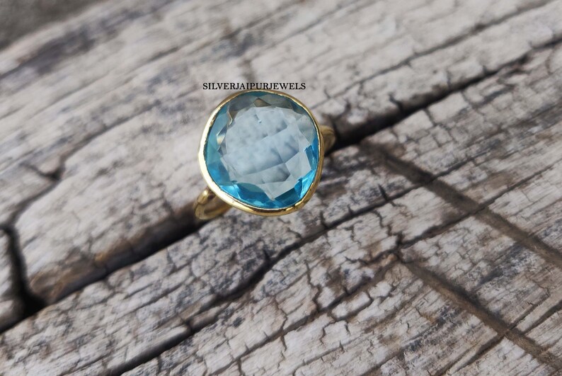 May include: A gold ring with a large, light blue gemstone. The gemstone is irregularly shaped and set in a gold bezel. The ring is on a weathered wooden surface. The text "SILVERJAIPURJEWELS" is visible.