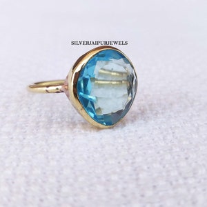 May include: A gold-colored ring with a large, light blue gemstone. The gemstone is faceted and set in a gold bezel. The ring is on a white surface. The text "SILVERJAIPURJEWELS" is visible.