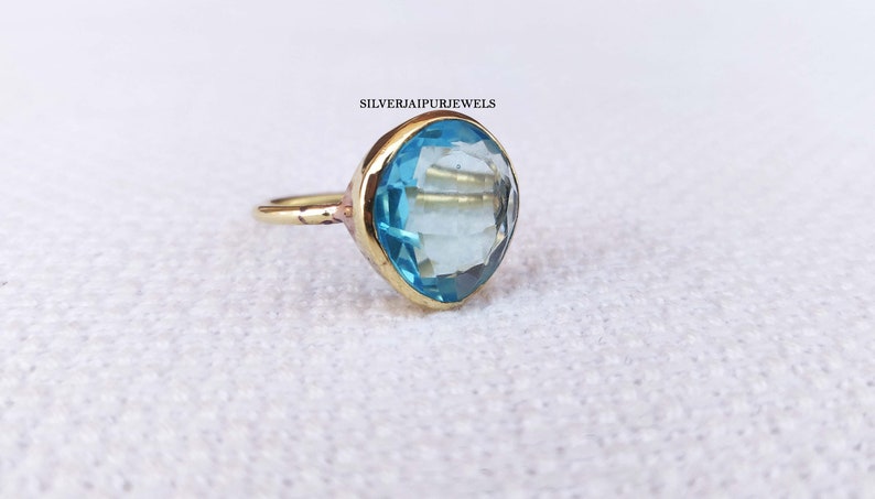 May include: A gold-colored ring with a large, light blue gemstone. The gemstone is faceted and set in a gold bezel. The ring is on a white surface. The text "SILVERJAIPURJEWELS" is visible.