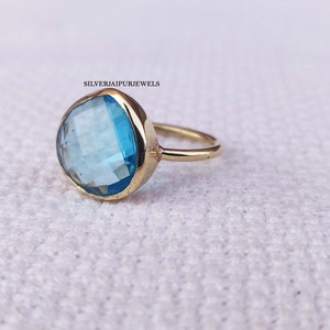 May include: A gold ring with a large, faceted, light blue gemstone. The ring is on a white, textured surface.