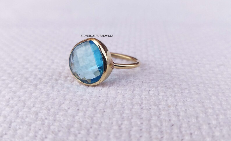 May include: A gold ring with a large, faceted, light blue gemstone. The ring is on a white, textured surface.