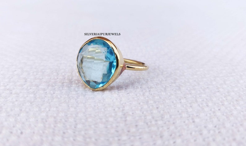 May include: A gold ring featuring a large, light blue gemstone with a faceted cut. The ring has a simple band and a bezel setting. The text "SILVERJAIPURJEWELS" is visible in the upper left corner.