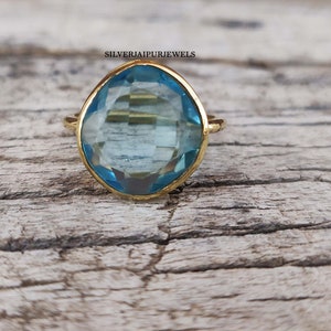 May include: A gold-colored ring with a large, light blue gemstone. The gemstone has a faceted cut and is set in a simple, elegant band. The ring is displayed on a weathered wooden surface.