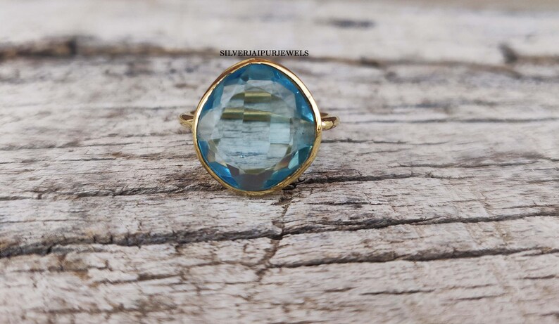 May include: A gold-colored ring with a large, light blue gemstone. The gemstone has a faceted cut and is set in a simple, elegant band. The ring is displayed on a weathered wooden surface.