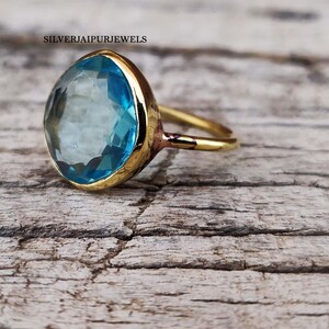 May include: A gold-colored ring with a large, light blue gemstone. The gemstone is oval-shaped and set in a gold bezel. The ring is on a weathered, light brown wooden surface. The text "SILVERJAIPURJEWELS" is visible.
