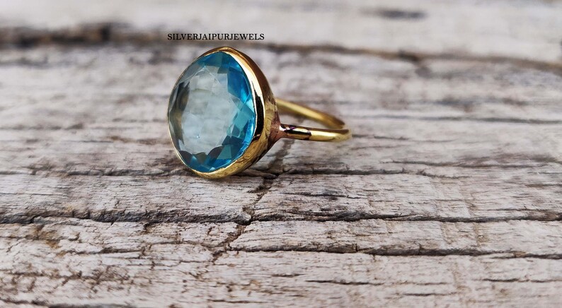 May include: A gold-colored ring with a large, light blue gemstone. The gemstone is oval-shaped and set in a gold bezel. The ring is on a weathered, light brown wooden surface. The text "SILVERJAIPURJEWELS" is visible.