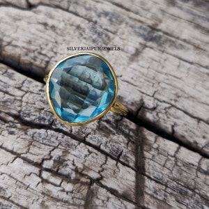 May include: A gold-toned ring featuring a large, faceted, light blue gemstone. The gemstone is set in a gold bezel and has a unique, irregular shape. The ring is displayed on a rustic, weathered wooden surface.
