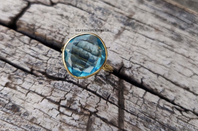 May include: A gold-toned ring featuring a large, faceted, light blue gemstone. The gemstone is set in a gold bezel and has a unique, irregular shape. The ring is displayed on a rustic, weathered wooden surface.