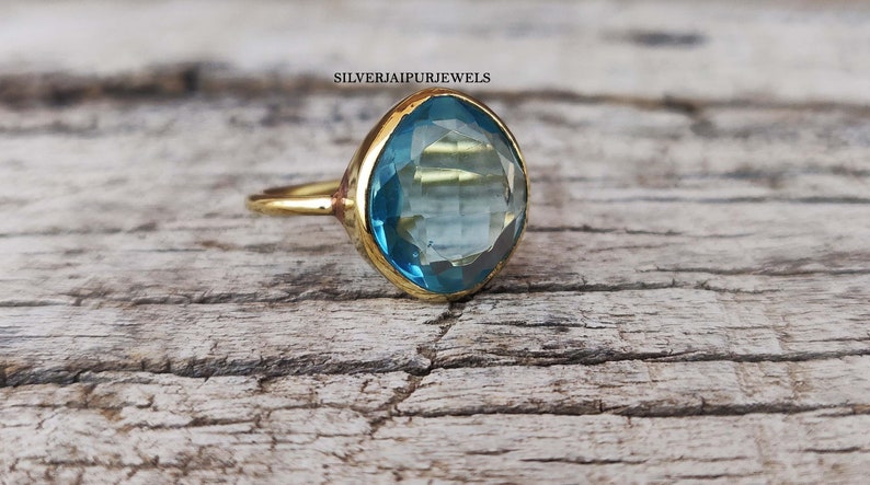 May include: A gold-toned ring with a large, light blue gemstone. The gemstone is a faceted, elongated cushion cut. The ring is set on a wooden surface. The text "SILVERJAIPURJEWELS" is visible.