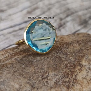 May include: A gold ring with a large, light blue gemstone. The gemstone is cushion-cut and set in a gold bezel. The ring is displayed on a textured, brown surface. The text "SILVERJAIPURJEWELS" is visible.