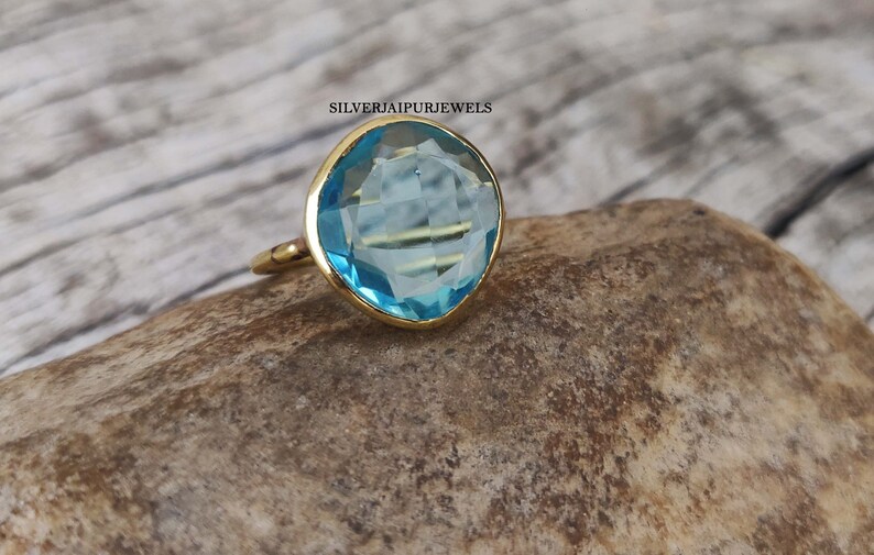 May include: A gold ring with a large, light blue gemstone. The gemstone is cushion-cut and set in a gold bezel. The ring is displayed on a textured, brown surface. The text "SILVERJAIPURJEWELS" is visible.