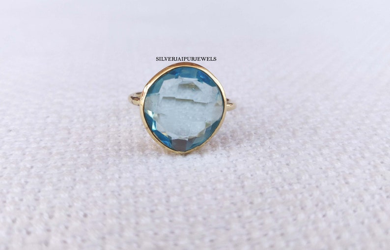 May include: A gold ring with a large, light blue gemstone. The gemstone is cushion-cut and set in a gold bezel. The ring is on a white, textured surface. The text "SILVERJAIPURJEWELS" is visible.