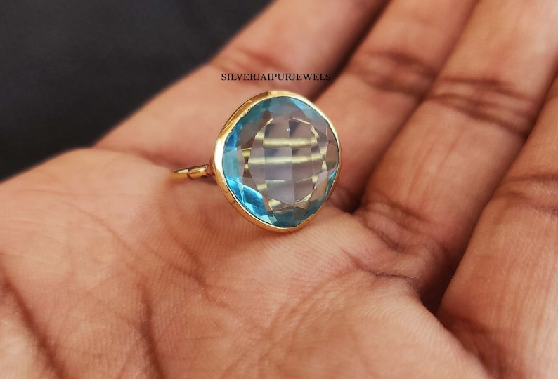 May include: A gold ring featuring a large, faceted, light blue gemstone. The gemstone is cushion-cut and set in a gold bezel. The ring is displayed on a hand, showcasing its color and design.