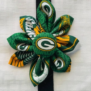 May include: Green and yellow fabric flower with a green "G" in the center. The flower is attached to a black elastic band.