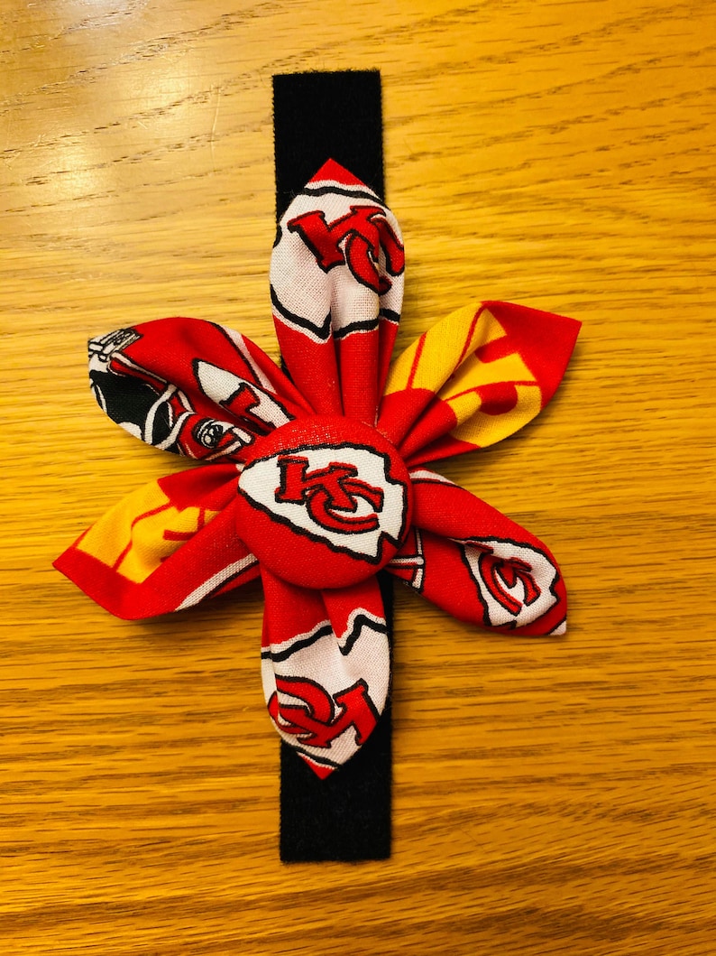 May include: A red, white, and yellow fabric flower with a red button in the center. The button has the Kansas City Chiefs logo on it. The flower is attached to a black elastic headband.
