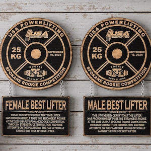 May include: Two circular wooden plaques shaped like 25 kg weight plates, labeled USA Powerlifting Spokane Rookie Competition September 19, 2026. Below each, a rectangular plaque hangs by chains, reading Female Best Lifter and Male Best Lifter.