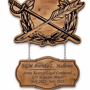 May include: A two-part wooden wall plaque featuring a top piece with a carved sword and quill crossed over a laurel wreath. A smaller rectangular wooden sign hangs below by two silver chains, engraved with SGM Brenda L. Hallows, Army Reserve Legal Command, G-3 Sergeant Major, July 2021-July 2023.