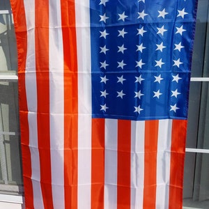 May include: A red, white, and blue American flag with 50 white stars on a blue field. The flag is hanging from a window.