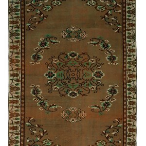 Puede incluir: Una alfombra rectangular de estilo vintage con un campo central marrón y intrincados diseños florales. La alfombra presenta un borde con diseños geométricos repetidos y motivos florales en tonos verdes, marrones y crema. Las dimensiones de la alfombra no son visibles.