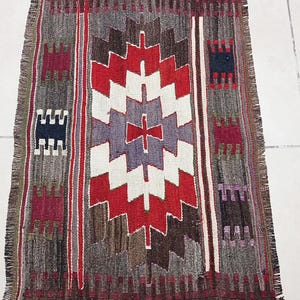 May include: A vintage rug with a geometric pattern in shades of red, white, brown, and purple. The rug has a fringe border and is woven with a traditional design.