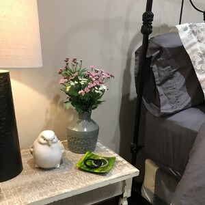 Puede incluir: Una mesita de noche de madera blanca con una figurita de pájaro de cerámica blanca, un plato de cerámica verde con pulseras negras y un jarrón de cerámica gris con flores rosas y blancas.