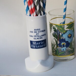 Può includere: Un contenitore in ceramica bianca con un'etichetta Marie Brizard contiene cannucce di carta colorate. Accanto c'è un bicchiere con disegni floreali blu e una cannuccia a righe blu e bianche. Il testo sul contenitore recita "Avec Jus de Citron et Eau Pétillante Glacée".